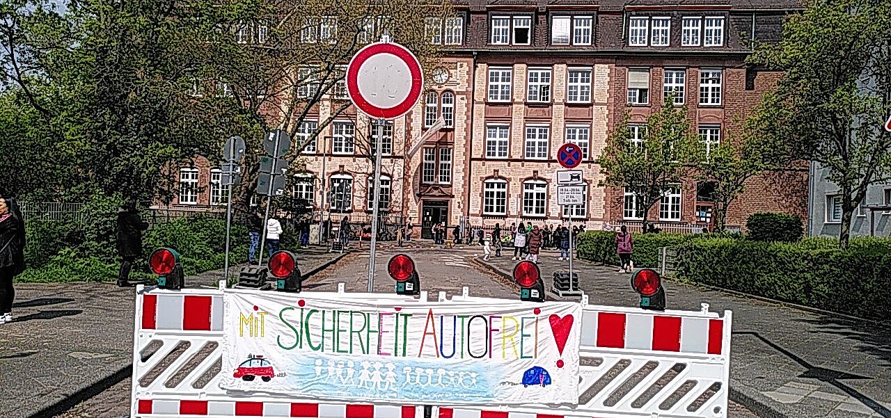 Das Bild zeigt die Sperre an der Schulstraße mit einem von Kindern gemalten Transparent mit der Aufschrift 'Mit Sicherheit Autofrei'