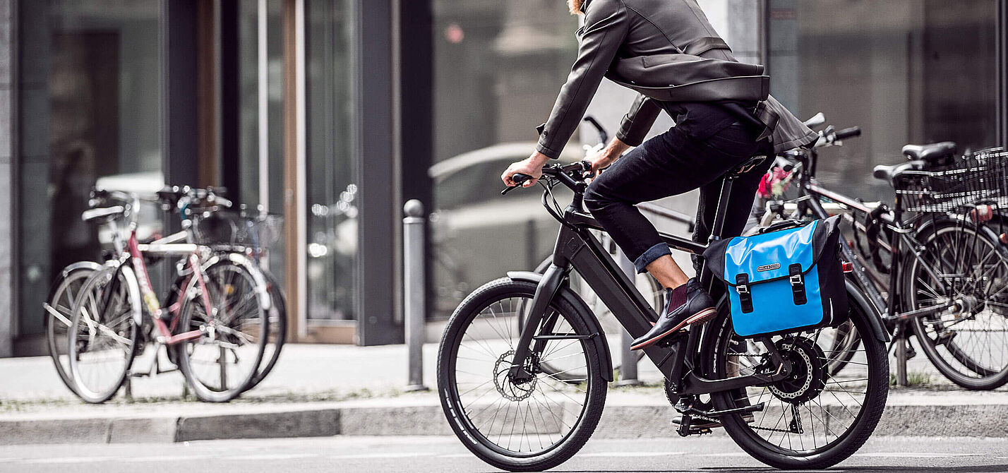Ein Mann mit Bart und weißem Helm fährt auf einem modernen Elektrofahrrad durch eine städtische Straße. Er trägt Alltagskleidung (Jacke und Jeans) und hat eine leuchtend blaue Packtasche am Gepäckträger befestigt. Im Hintergrund sind Gebäude mit großen Glasfronten und parkende Fahrräder unscharf zu erkennen.