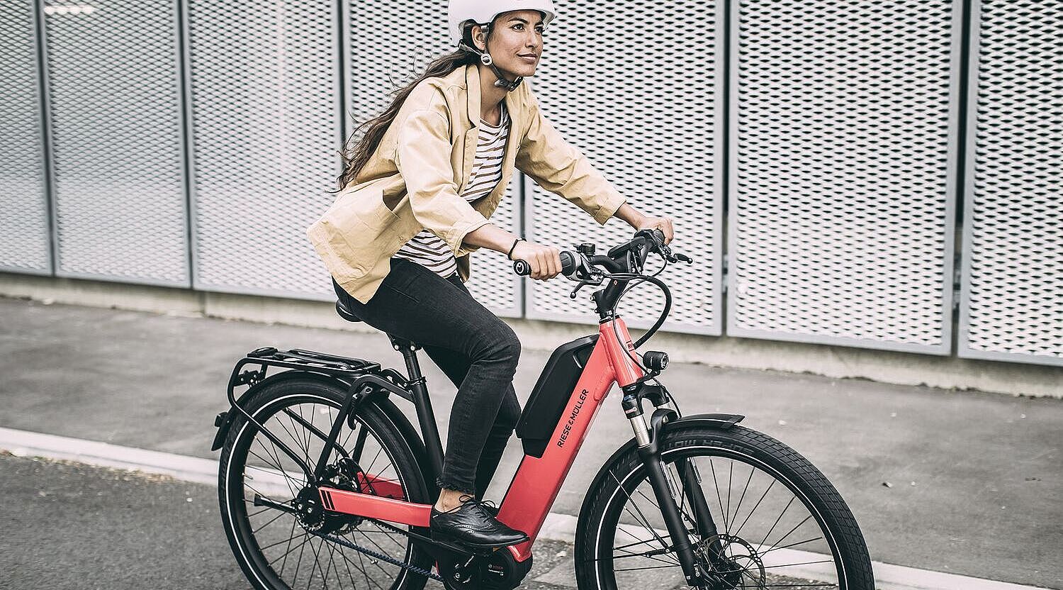 Eine Frau fährt auf einem roten Elektrorad auf Asphalt, im Hintergrund graue, gelochte Metallwände. 
